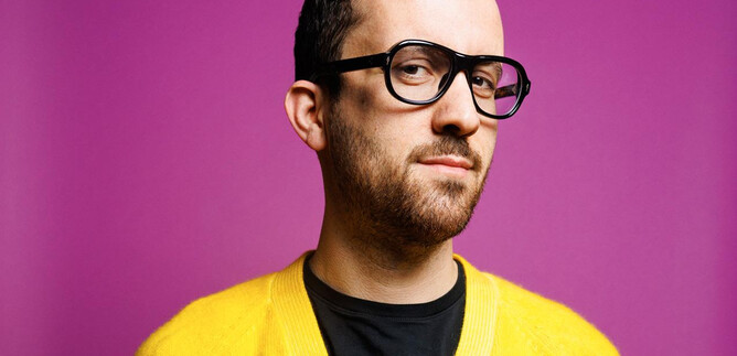 A man with glasses and a colorful sweater is standing in front of a pink background. He is looking thoughtfully at the camera. | © Peter Rigaud
