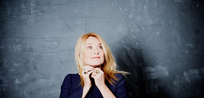 A woman with blonde hair gazes thoughtfully upwards. She is wearing a dark blue top in front of a gray wall. | © Julia Wesely