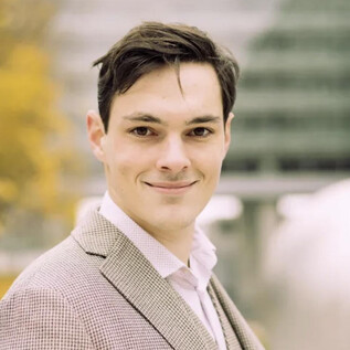 A young man in an elegant suit smiles warmly at the camera. In the background, modern buildings and autumn trees can be seen. | © Lukas Beck
