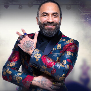 A man in a colorful, floral patterned suit smiles and makes a hand gesture. In the background, blurred silhouettes can be seen, conveying a fighting mood. | © @hypnox-akademie