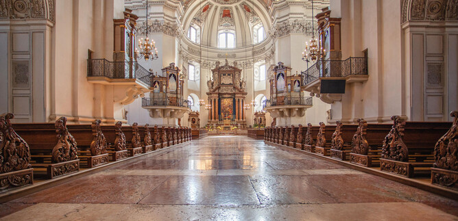 An impressive church with a high vault and ornate decorations. The wooden pews lead to the magnificent altar area. | © Erzdiözese Salzburg