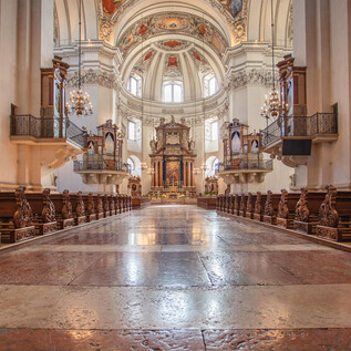 An impressive church with a high vault and ornate decorations. The wooden pews lead to the magnificent altar area. | © Erzdiözese Salzburg