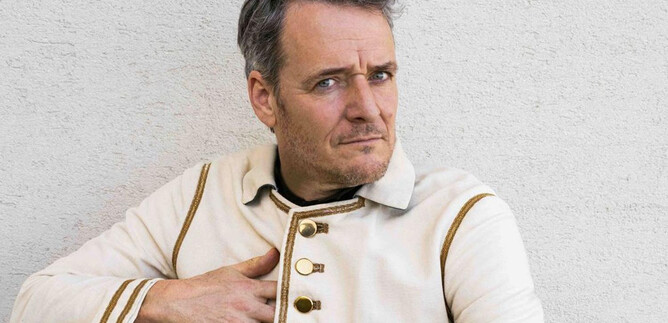 A man in a striking white jacket with golden buttons sits comfortably. He has short gray hair and is looking directly at the camera. | © Paul Vincent Schütz