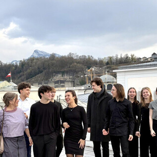 A group of people stands on a rooftop overlooking an urban landscape. They are dressed in elegant, dark clothing and appear cheerful in their shared environment. | © Universität Mozarteum