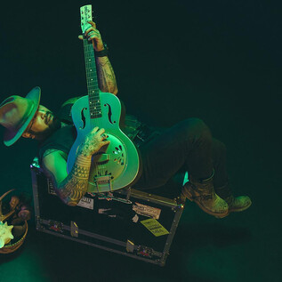 A musician is sitting on a box and playing the guitar. He is wearing a hat and is surrounded by green light. | © Rockhouse Salzburg