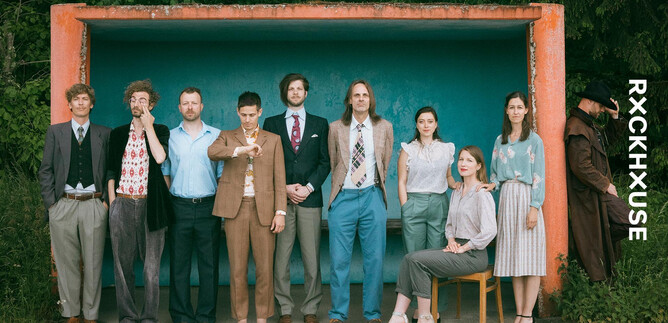A group of nine people in formal attire stands in front of a colorful background. They are posing in different stances and showcasing a diverse use of colors and styles. | © Sonja Herpich