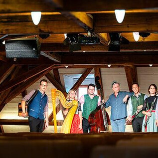 A group of musicians stands in a room with wooden ceilings and warm light. They have various instruments and are wearing traditional clothing. | © Constanze Passin