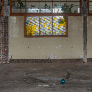 An empty, industrial-looking interior with a round, metallic object and a green sphere on the floor. Behind a colorful, tasteful window, plants are visible. | © Sarah Pichlkostner