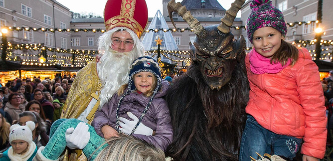 A festive scene with two children dressed up as Santa Claus and a Krampus. In the background, a bustling Christmas market atmosphere can be seen. | © Salzburger Christkindlmarkt
