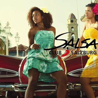 Two women pose in colorful dresses next to a classic red car. In the background, buildings and palm trees can be seen, creating a vibrant atmosphere. | © Jazzit