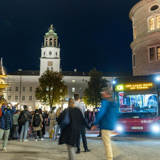 A lively nighttime scene with a bus stopping at a station. In the background, historic buildings and a crowd of people can be seen. | © wildbild