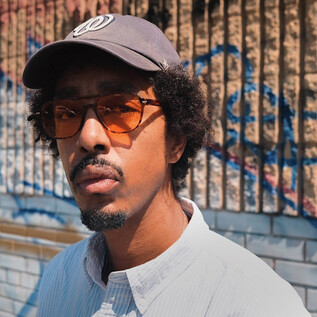 Muž stojí před barevnou zdí s graffiti. Na sobě má kšiltovku a sluneční brýle a má kudrnaté vlasy. | © Rockhouse Salzburg