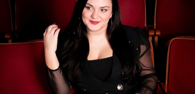 A stylish woman is sitting in a theater with red seats. She is smiling and wearing an elegant black outfit. | © Amer productions