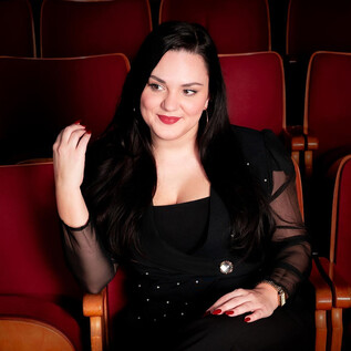 A stylish woman is sitting in a theater with red seats. She is smiling and wearing an elegant black outfit. | © Amer productions