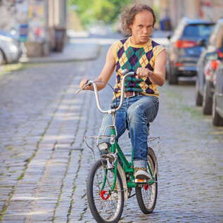 A man rides a green bicycle along the cobbled street. The surroundings are urban with cars parked at the side of the road. | © Amac Garbe