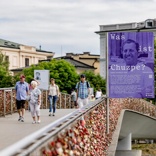 A bridge with several people walking. On the sides, there are large posters with information and questions affixed. | © wildbild