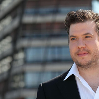 A man in a suit stands in front of a modern building. He has a confident expression and looks at the camera. | © christian-kargl