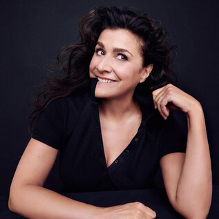 A woman with curly hair sits smiling at a table. The background is dark, which enhances her friendly demeanor. | © Kristian Schuller_Decca