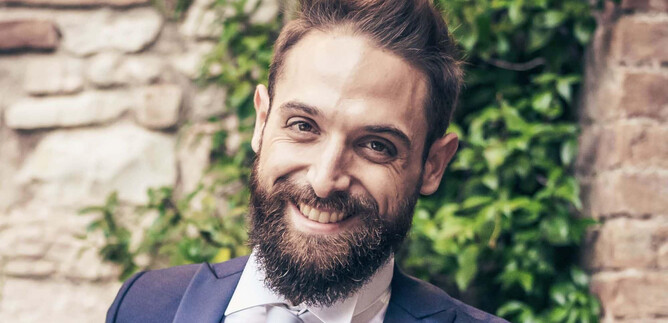 A smiling man in an elegant suit stands in front of a stone wall with green plants. He has a fuller beard and appears friendly and confident. | © Ariosi Artits Management