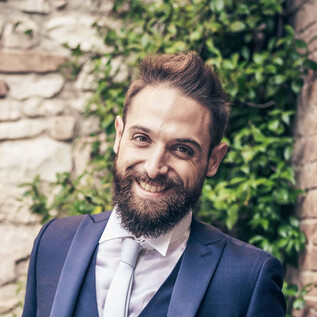 A smiling man in an elegant suit stands in front of a stone wall with green plants. He has a fuller beard and appears friendly and confident. | © Ariosi Artits Management