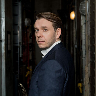 A man in a suit is looking over his shoulder in a narrow, illuminated corridor. The background is dark and industrial. | © Edouard Brane