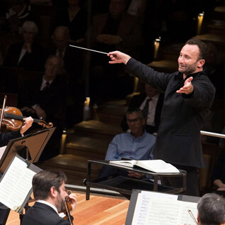 An orchestra conductor passionately leads a performance with expressive gestures. In the foreground, a violinist plays while the audience listens attentively. | © Monika Rittershaus