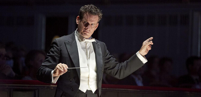 A conductor leads an orchestra with great passion. In the background, spectators can be seen during a concert. | © Michael Poehn