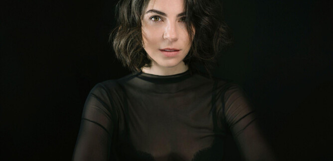 A woman with short, curly hair is wearing a black, transparent top. She is looking directly into the camera against a dark background. | © Chris Gonz
