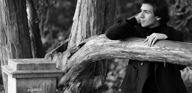 A thoughtful young man leans against a tree trunk outdoors. The image composition is in black and white. | © Sasha Gusov