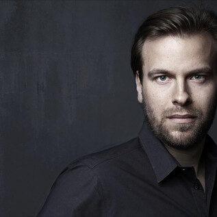A portrait of a young man with short dark hair and a beard. He is wearing a black shirt and looking directly at the camera. | © Gisela Schenker