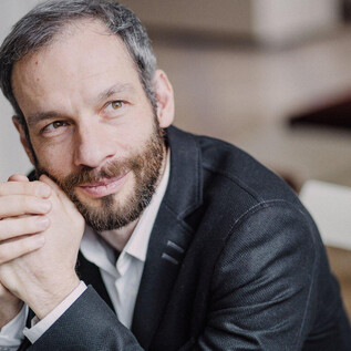 A man with short hair and a beard sits thoughtfully. He is wearing a black suit and looking kindly at the camera. | © Marco Borggreve