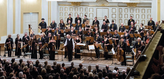 An orchestra in full force is playing on a stage. The audience in the background is applauding enthusiastically. | © Albert Moser