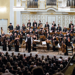 Un orchestre complet joue sur une scène. Le public en arrière-plan applaudit avec enthousiasme. | © Albert Moser