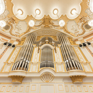A large, magnificent organ with many pipes in a festively designed room. The walls are painted in soft colors and adorned with golden embellishments. | © Wolfgang Lienbacher