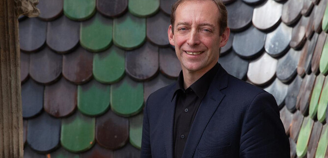 A man smiles in front of a colorful shingle facade. He is wearing a dark suit and has a friendly expression. | © Verein "Unser Stephansdom"