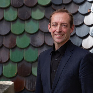 A man smiles in front of a colorful shingle facade. He is wearing a dark suit and has a friendly expression. | © Verein "Unser Stephansdom"