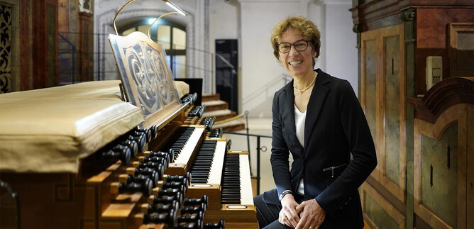 A person is sitting at an organ and smiling. In the background, intricately designed architecture can be seen. | © www.salzburger-dom.at