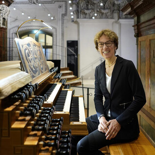 A person is sitting at an organ and smiling. In the background, intricately designed architecture can be seen. | © www.salzburger-dom.at