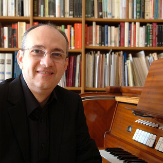 A man sits smiling next to a musical instrument in a room with bookshelves in the background. He is wearing a black shirt and glasses. | © Roberto Marini