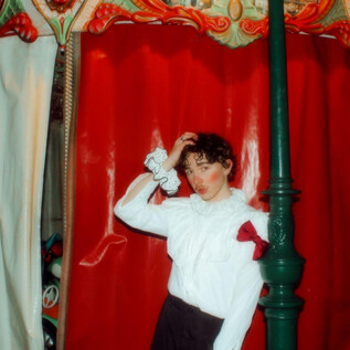 A young model stands in front of a red curtain and poses. She is wearing a white top with a red bow and has a playful demeanor. | © Michelle Rassnitzer