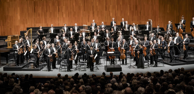 A large orchestra stands on stage and plays a concert. Many listeners can be seen in the audience. | © Stephan Rabold