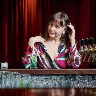 A woman is sitting at a bar and watching attentively. In the background, glasses and a counter with taps can be seen. | © Foto Flausen