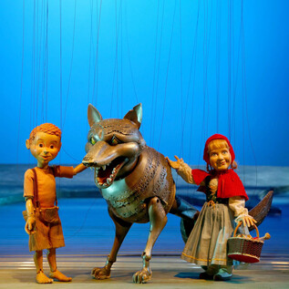 A boy doll, a girl doll in a red cloak, and a wolf doll stand together. The background is blue, creating an imaginative atmosphere. | © Salzburger Marionettentheater