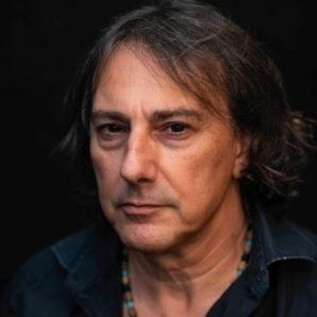 A man with long, dark hair and a serious expression stands in front of a dark background. He is wearing a black shirt and a pearl necklace. | © derkonzertfotograf
