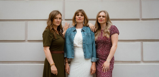 Three women are smiling in front of a bright wall. They are wearing different dresses and have a modern, stylish look. | © Laura Edl