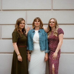 Three women are smiling in front of a bright wall. They are wearing different dresses and have a modern, stylish look. | © Laura Edl