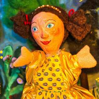 A colorful doll with curly hair and yellow clothing. In the background, cheerful colors and a fairy tale landscape can be seen. | © Hannes Öhlböck