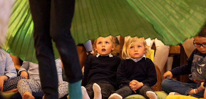 Two children are sitting excitedly on the floor watching a performance. In the background, a colorful curtain can be seen. | © Erika Mayer