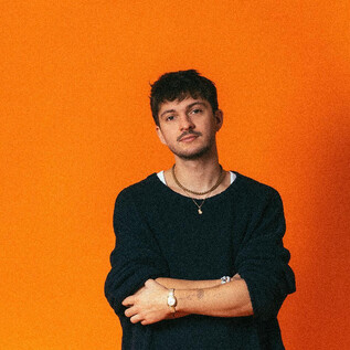 A young man is standing in front of an orange background. He is wearing a black top and has a relaxed pose. | © Mario Wallner