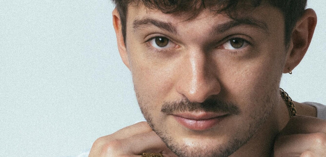A young man with short curly hair and a light beard. He is wearing a white T-shirt and adorns himself with several pieces of jewelry. | © Mario Wallner